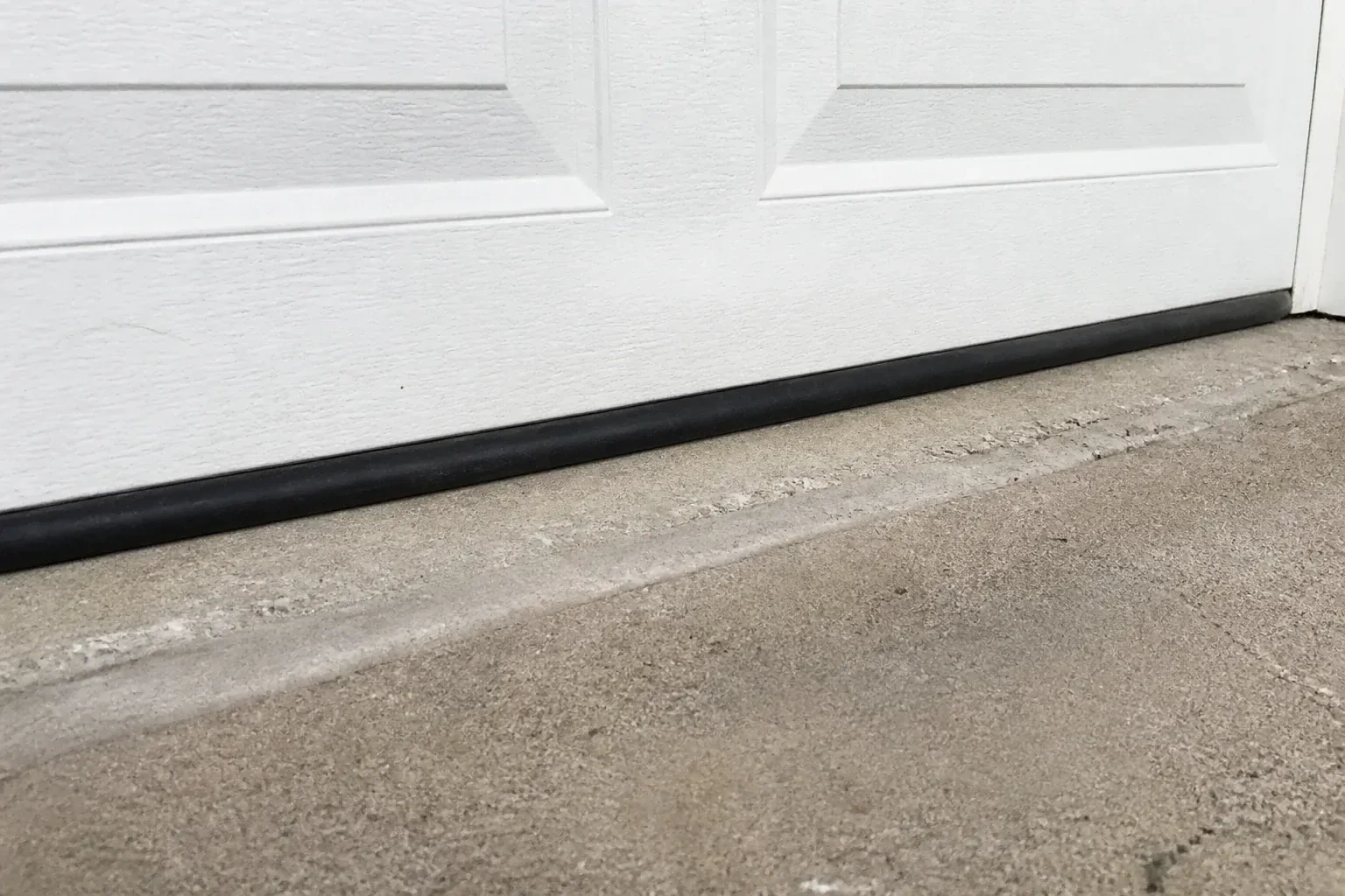 Black bottom seal along a closed white garage door at a concrete threshold. — Garage Door Bottom Seal Replacement, Hamilton