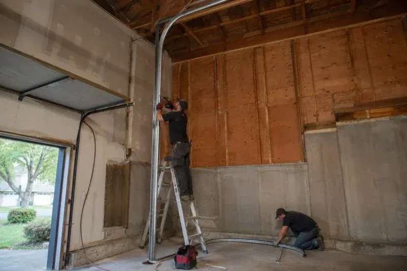 One person on a stepladder working beside a tall curved garage-door track while another kneels beside loose track sections in an unfinished garage. — High-Lift Garage Door Conversion, Hamilton